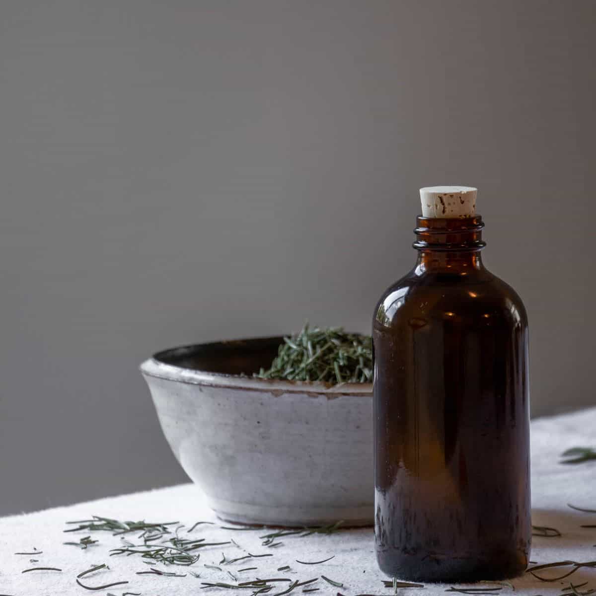 A bottle of rosemary oil next o a bowl of dried rosemary leaves